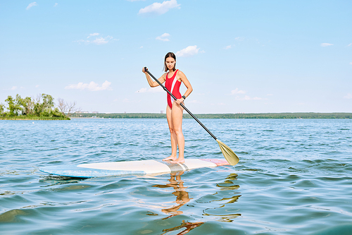 Young pretty brunette sportswoman in red swimsuit standing on surfboard and using paddle while floating on water against sky and horizon