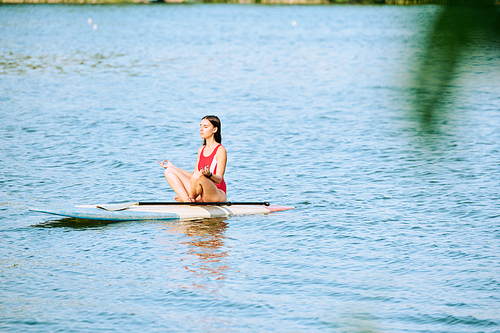 Young serene female in red swimsuit sitting on surfboard in pose of lotus and floating on water while enjoying vacation at summer resort