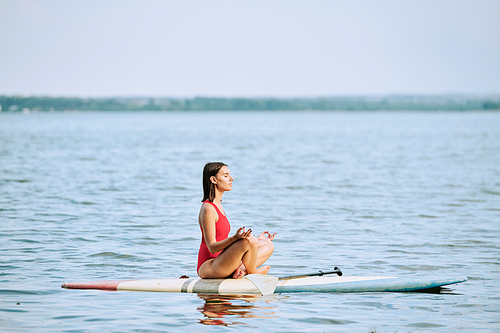 Young sportswoman in red swimsuit practicing yoga while sitting on surfboard in pose of lotus and floating on water at resort