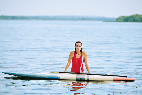 Happy young brunette woman in red swimsuit standing in water by surfboard while enjoying solitude and summer vacation at resort