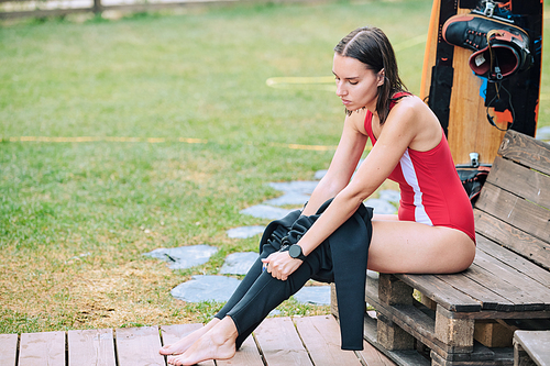 Pretty brunette sportswoman with dark wet hair putting on black leggins while sitting on wooden bench before surfboard training