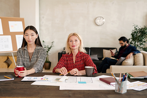 Young Asian female office worker and her blond mature colleague having coffee while sitting by table with many papers against businessman