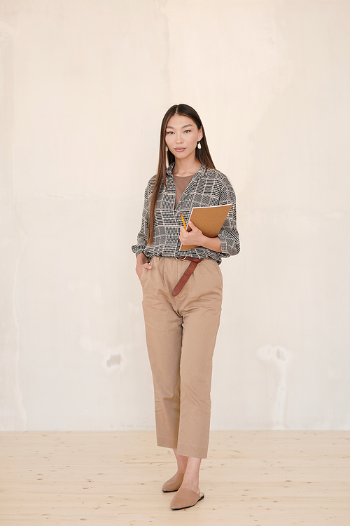 Young businesswoman with dark long hair holding notebook and pencil while standing in front of c amera against white wall in studio