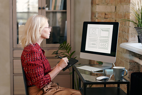 Blond mature female mobile application developer with tablet and stylus sitting by table in front of computer monitor and drawing sketches