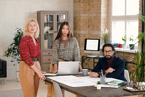 Group of two elegant females and young bearded man looking at you while making sketches of new mobile application by workplace