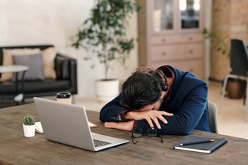 Young tired or sick businessman in formalwear keeping his head on arms while napping on wooden table in front of laptop after work in office