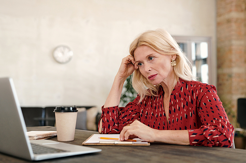Serious mature blond businesswoman sitting in front of laptop and looking at online data on display during network in office environment