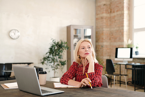 Pensive mature blond businesswoman sitting in front of laptop and contemplating about some working points in office environment