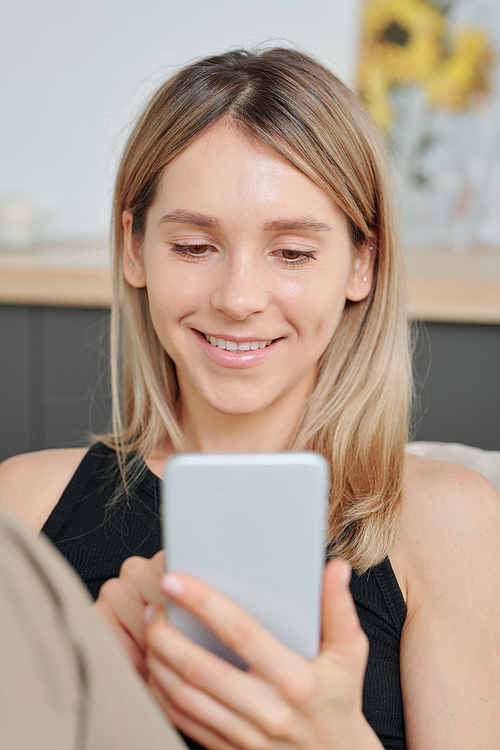 Young cheerful female with long blond hair looking at her friend on smartphone screen with smile during communication in video chat