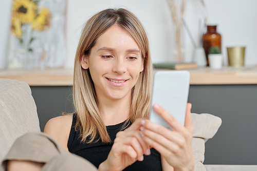 Cheerful millennial with long blond hair looking at smartphone screen while scrolling through online videos or photos of friends