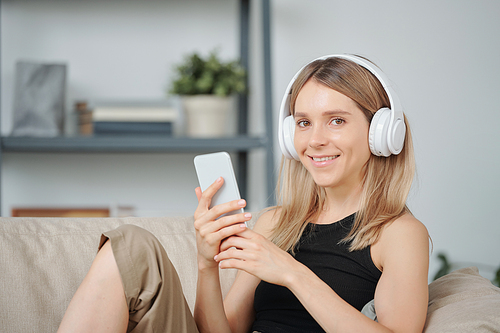 Happy young female with toothy smile listening to music in headphones and looking at you while relaxing on couch in home environment