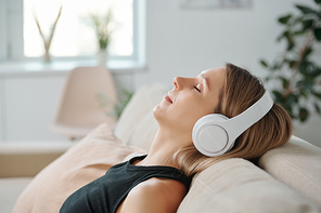 Side view of young relaxed woman in headphones lying on back of couch and listening to relaxing music while enjoying home rest