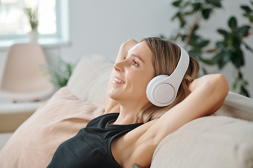 Side view of young blond smiling woman in headphones enjoying music while lying on back of couch in home environment and having rest