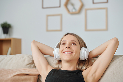 Young blond cheerful woman in headphones keeping hands behind head while lying on back of couch in home environment and having rest