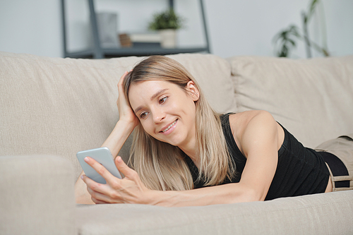 Pretty blond girl with smartphone lying on soft couch at leisure and watching video in social networks while relaxing at home