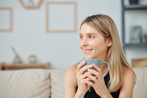 Contemporary young woman with toothy smile and long blond hair holding mug with hot drink while enjoying morning coffee