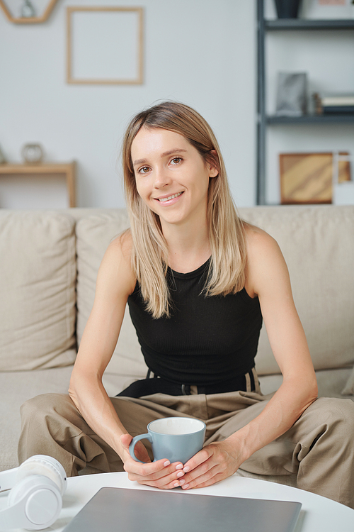 Happy blond female in casualwear looking at you with smile while sitting on couch in front of camera, having drink and enjoying time at home