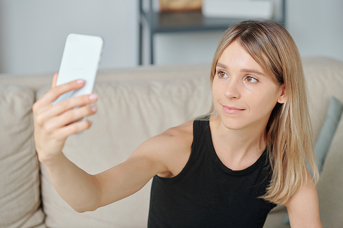 Young smiling blond female in black vest looking in smartphone camera while holding gadget in front of her face and making selfie on