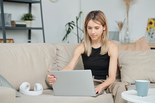 Contemporary young woman in tanktop and pants sitting on soft couch in living-room in front of laptop and going to watch movie at leisure