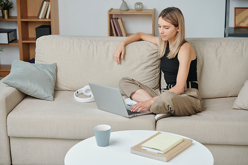 Contemporary young blond woman with crossed legs sitting on soft couch in living-room and using laptop while surfing in the net at leisure