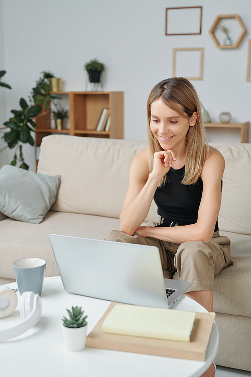 Young smiling female with long blond hair sitting on couch in front of table with laptop and looking at display during communication in video chat