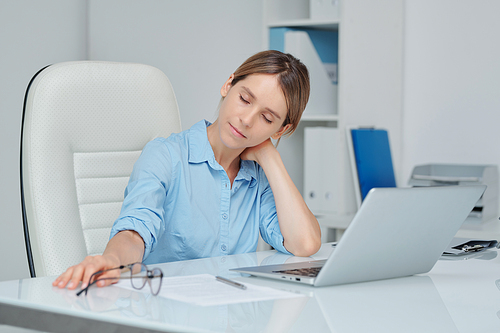Young tired businesswoman touching back of her neck while sitting by desk in front of laptop and trying to relax during short break