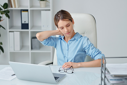 Young elegant businesswoman with tense feeling in neck touching or massaging the zone with discomfort or pain while sitting by desk