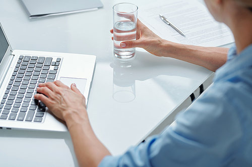 Hands of contemporary businesswoman in blue shirt sitting by desk in front of laptop, having glass of water and pushing buttons of keypad