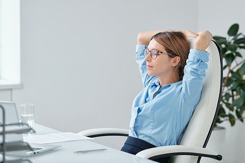 Young restful businesswoman in eyeglasses and blue shirt keeping hands behind head while relaxing in armchair and keeping her eyes closed