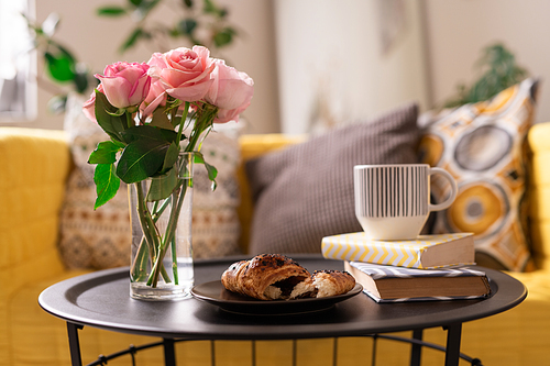 Bunch of fresh pink roses in glass of water, homemade croissant, cup of tea or coffee and two books on tray on background of couch