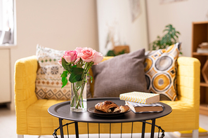 Two books, fresh croissant on plate and bunch of pink roses in glass of water on small table with couch on background in living-room