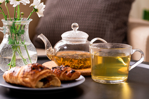 Teapot and cup with green herbal tea, fresh croissant on plate and bunch of flowers on table on background of pillow on armchair