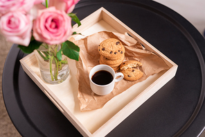 Low square carton box with cup of coffee and crispy cookies on paper, bunch of pink roses in glass on small black round table