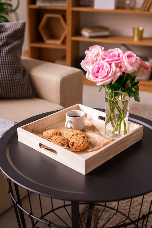 Square wooden box with bunch of fresh pink roses in glass, cup of coffee and cookies on small round table in home environment