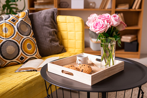 Home interior with pillows and open book on couch and small table with cookies, cup of coffee and bunch of pink roses in wooden box