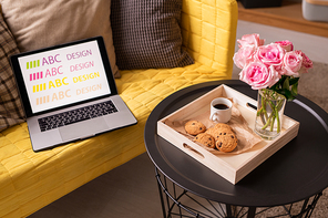 Laptop and cushions on yellow couch by small table with wooden box containing bunch of pink roses, drink and tasty cookies