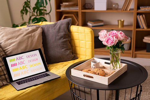 Small round black table with roses, cookies and cup of coffee standing by yellow sofa with pillows and laptop of designer in living-room