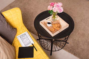 Bunch of pink roses, coffee and cookies in wooden box on small table by yellow couch with cushions and tablet with electronic sketch