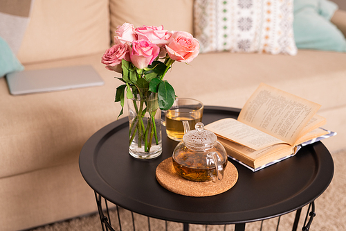 Bunch of pink roses in glass of water, open book, teapot and cup with green tea on small table by comfortable couch with cushions
