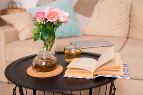 Small table with rose bunch in glass of water, green tea in teapot and cup and open book of stories against couch with pillows and tablet