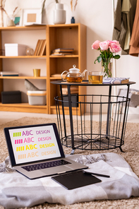 Laptop and pad with stylus on plaid by small table with herbal tea and pink roses standing on the floor with bookshelf on background