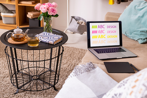 Small table with book, green tea and pink roses standing on soft rug by comfortable couch with pad, stylus and laptop of designer