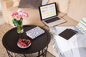 Red grapes on saucer, pink roses in glass of water and book on small table by couch with laptop, pad and stylus inside domestic room