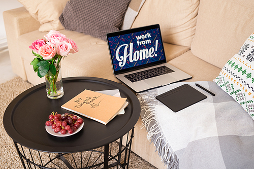 Small round table with bunch of roses, fresh red grapes on plate and book of sketches standing by couch with laptop and pad of designer