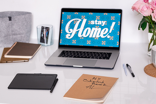 Laptop with stay home announcement on display, pad with stylus, sketchbook, pens, notebooks and roses on workplace of designer