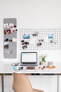 Chair by desk with laptop, smartphone, notebook with pen, mug with drink, flowers in jug and cosmetic products with photos above on wall