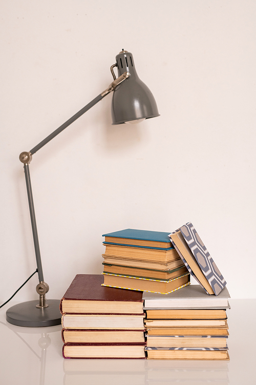Lamp over stacks of books and manuals on table or workplace of student of college or school against white wall in studio