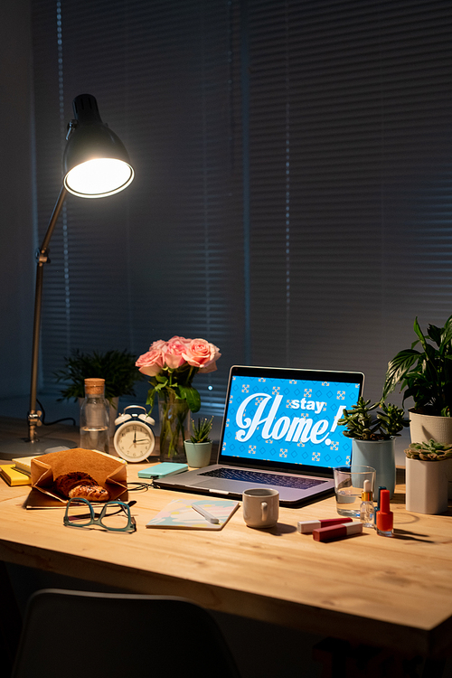 Lamp over wooden table with flowers, domestic plants, laptop, snack, drink, alarm clock, cosmetic products, eyeglasses and copybook in dark room