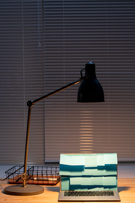 Light of lamp over laptop with blue notepapers on display and basket with highlighter near by on wooden table by window with jalousie