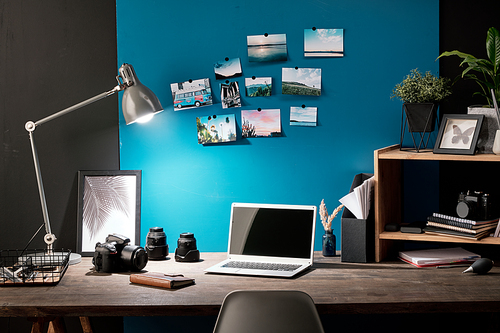 Workplace of photographer with photos of nature on blue board in front of table with photocamera, laptop, lamp, notebook and other stuff